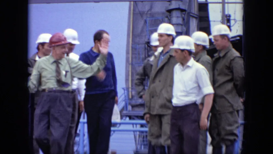 1971 Moscow frame of a supervisor in a red hard hat gesturing to workers in white hard hats at an industrial site.