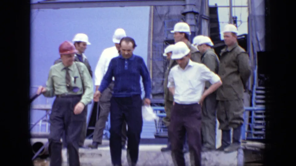 1971 Moscow home-movie frame showing a red-hard-hat supervisor and workers moving from a platform near industrial equipment.