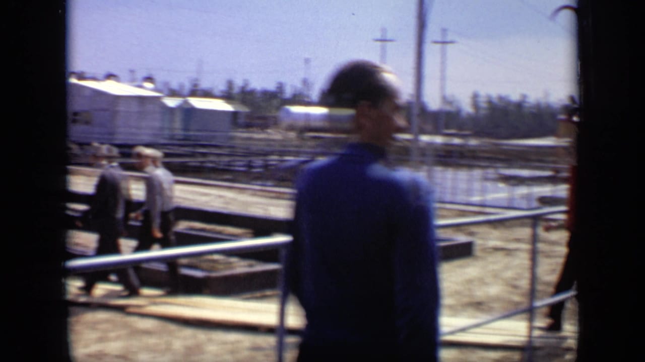 In 1971 Moscow footage, men in suits and shirts walk across planks near railings, muddy ground, and industrial structures.