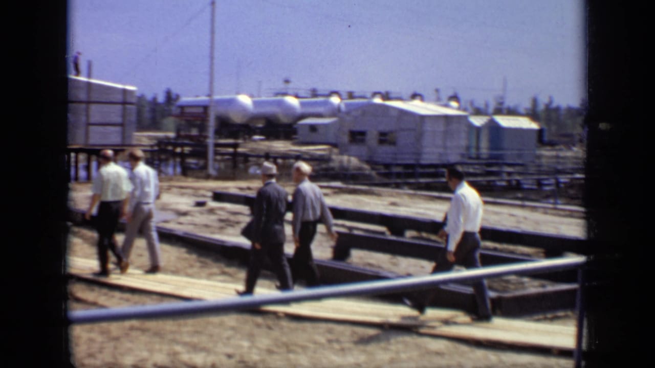 Men continue walking past industrial tanks and temporary buildings on a construction site.