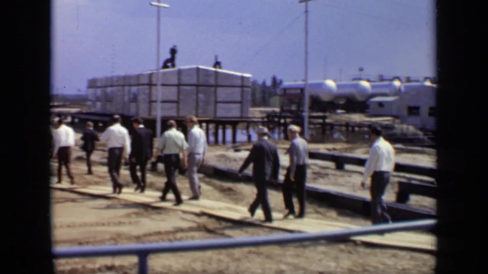 Several men in suits and shirts move across wooden planks at the Moscow construction site, with a raised framed structure and tanks visible nearby.