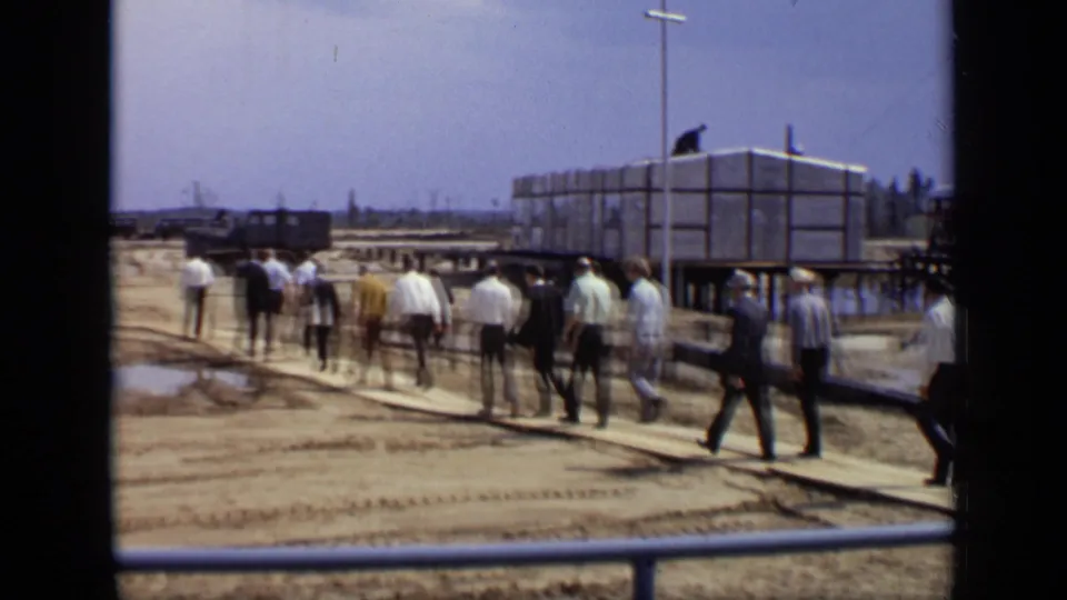 The group walks farther into the 1971 Moscow industrial site, crossing planks over muddy ground near temporary structures and utility poles.