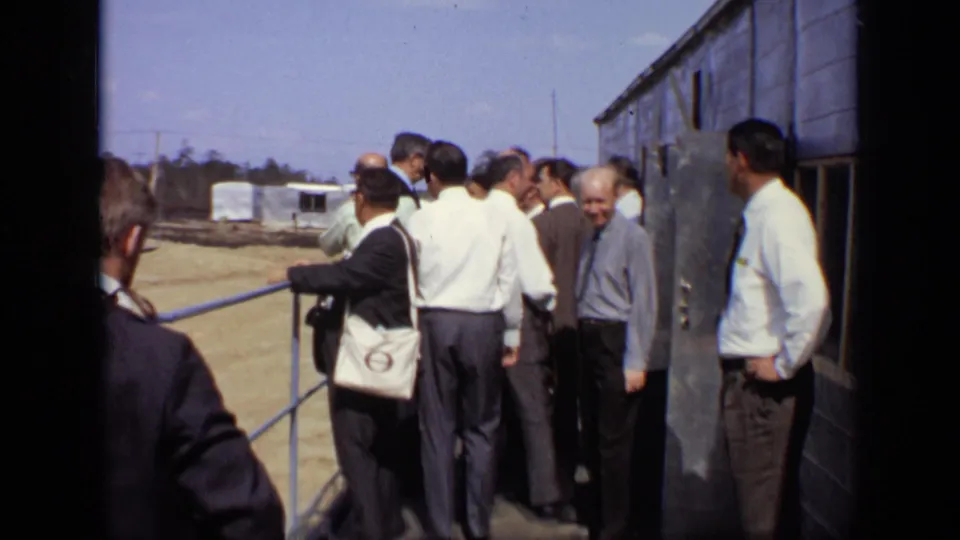 A group of adult men stands close together near a railing and industrial wall, with the construction site visible beyond.