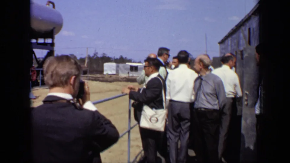 A man raises a camera while other visitors stand by the railing near storage equipment and an industrial building in the 1971 Moscow clip.