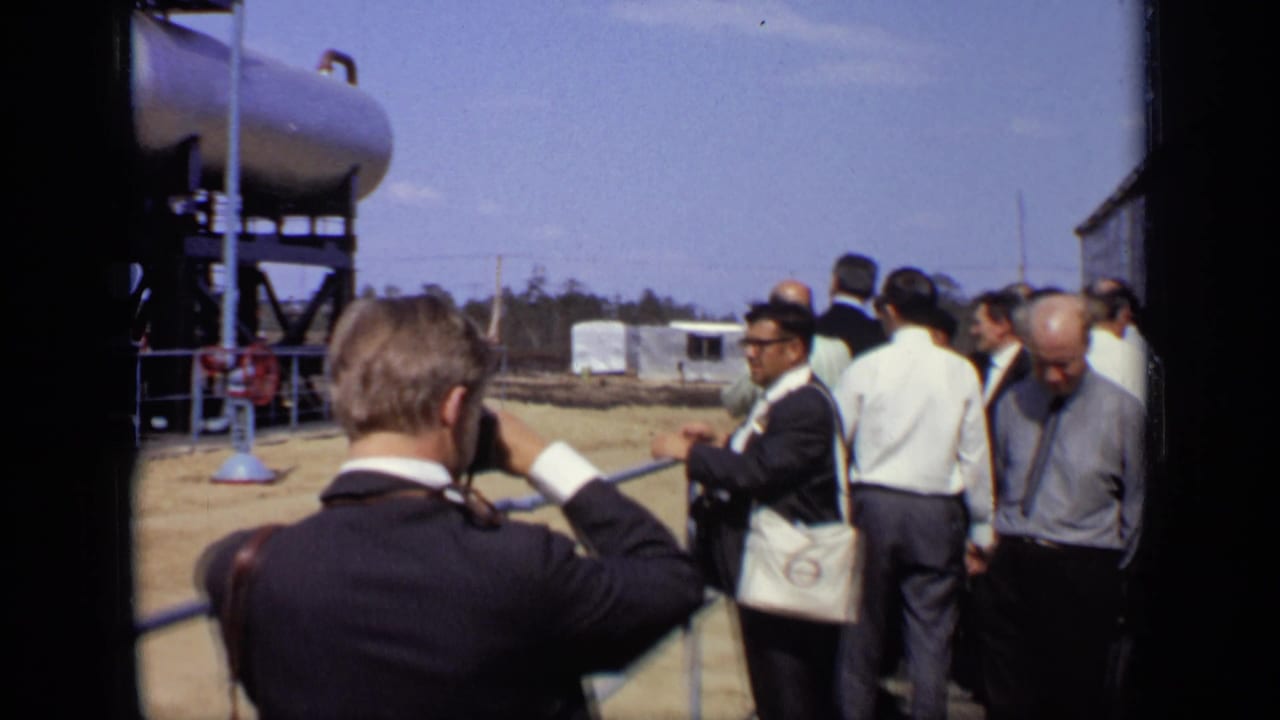 In 1971 Moscow footage, adult men in suits and shirts stand behind a railing while viewing large elevated silver tanks outdoors.