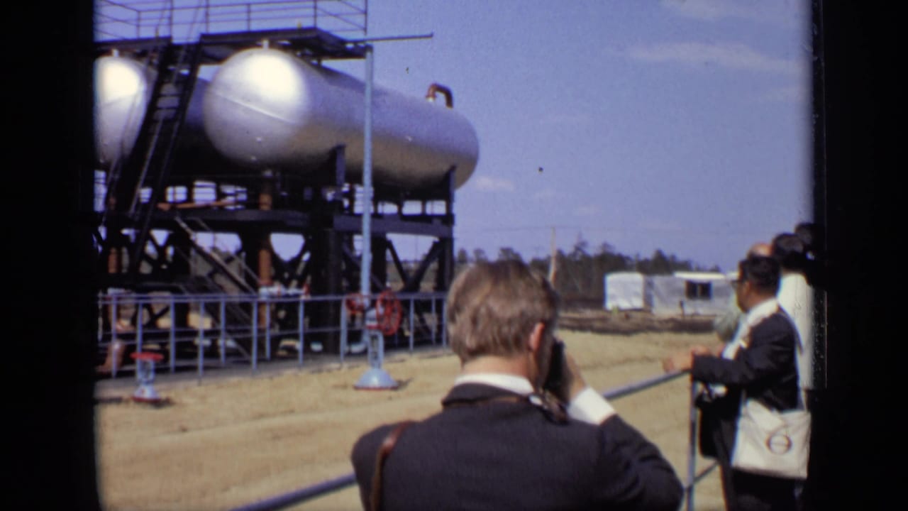 A man with a camera faces large elevated tanks as other visitors stand nearby.