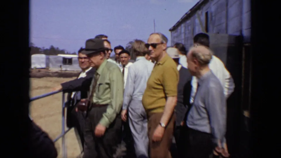 Adult men in period shirts, suits, hats, and sunglasses gather near a dark building wall and railing at the industrial site.