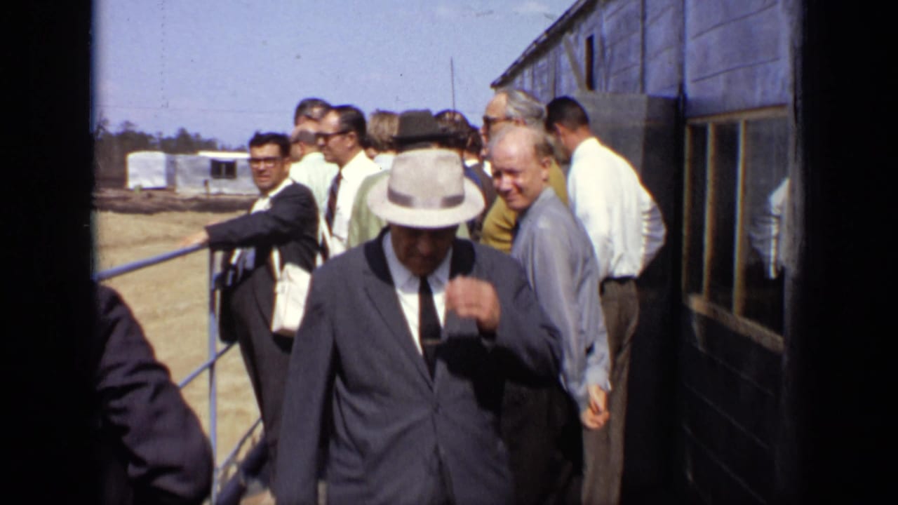 Adult men in suits leave a building, with one man in a fedora adjusting his hat.