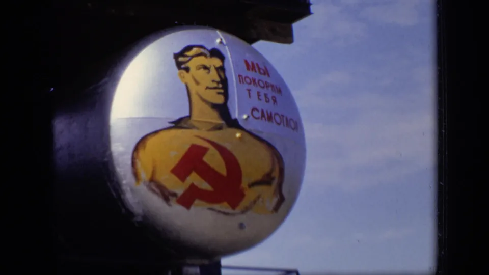 Close 1971 Moscow frame of a cylindrical tank end with a painted worker, hammer and sickle emblem, and Russian writing beside blue sky.