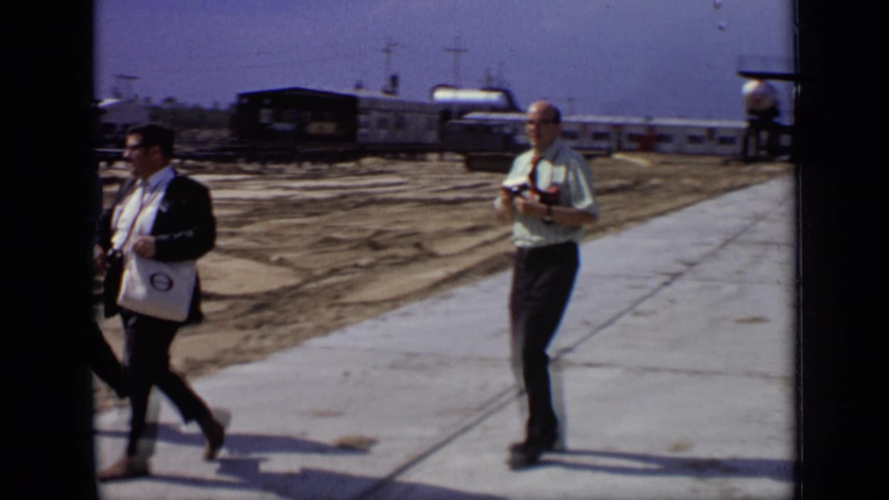 Two adult men walk through a Moscow industrial setting in 1971, one holding a camera as railcars and tanks sit in the background.