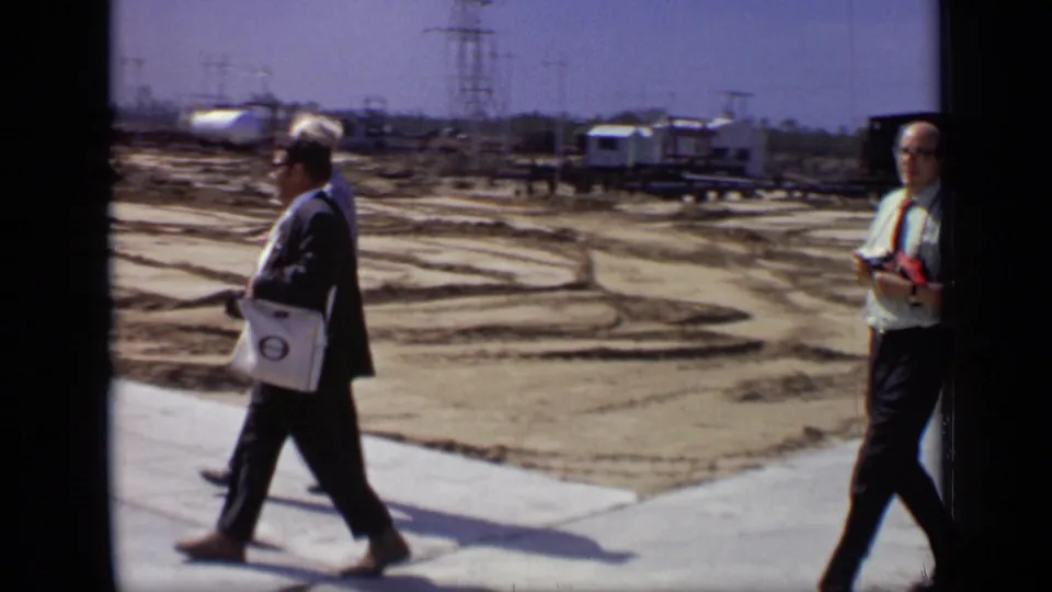 A suited man with a white shoulder bag walks through a 1971 Moscow industrial area while another man with a camera stands farther back.