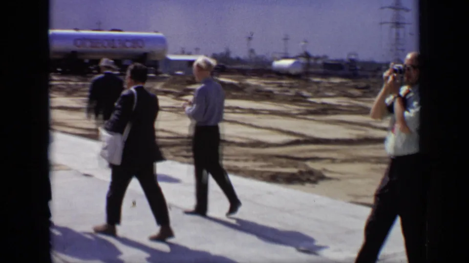 Three adult men move through the Moscow industrial site, with one raising a camera near tanks, railcars, and open ground.
