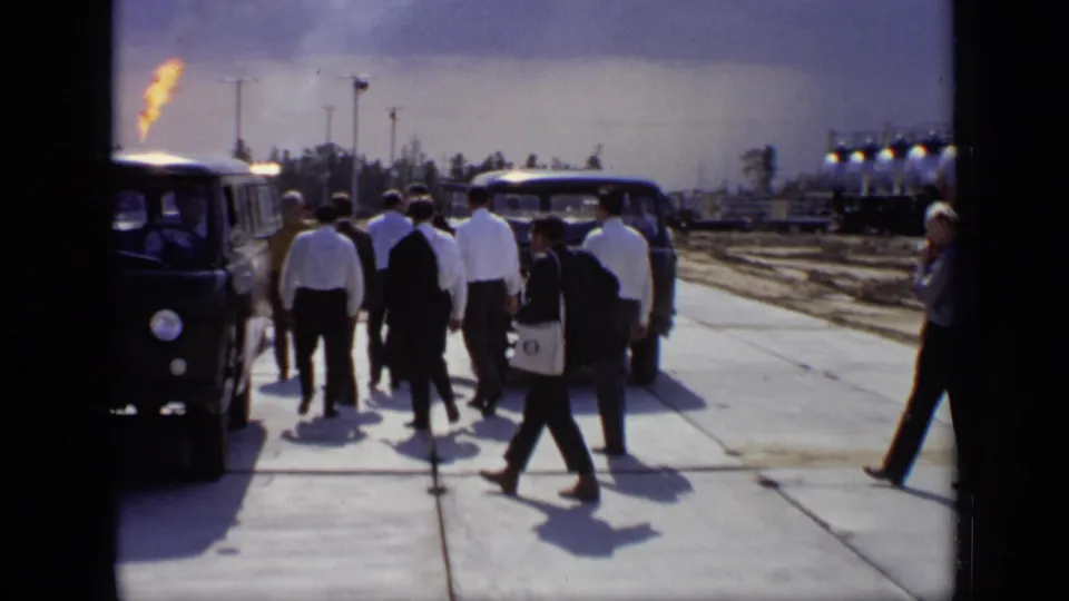 The group moves through the industrial site as a gas flare burns beyond parked utility vehicles and flammable tankers.