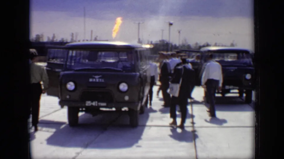 A dark utility van sits in the foreground while officials stand nearby and a gas flare burns over the industrial site.