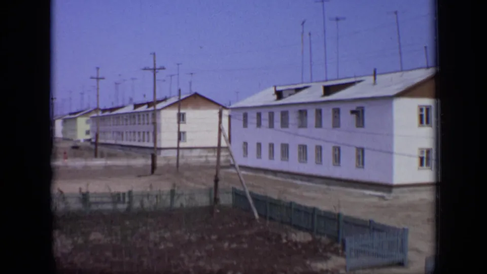 1971 Moscow 8mm frame showing simple white residential buildings, utility poles, and a fenced dusty street edge.