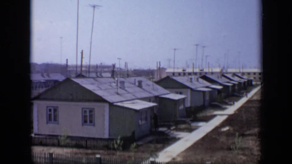 1971 Moscow train-view frame showing a row of small houses, a straight path, rooftop antennas, poles, and distant industrial buildings.