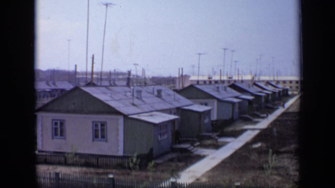 Similar view of small houses, walkway, open ground, poles, and distant low buildings.
