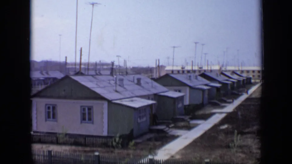 1971 Moscow home-movie frame showing rows of modest houses, a long paved walkway, utility poles, fencing, and distant buildings.