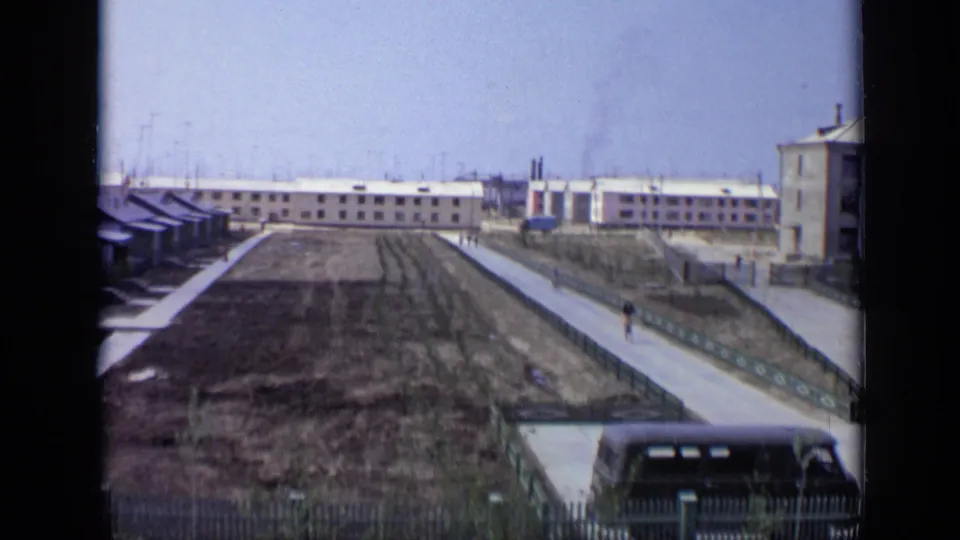 1971 Moscow residential area with open lots, paved paths, low buildings, a parked dark vehicle, and a distant pedestrian.