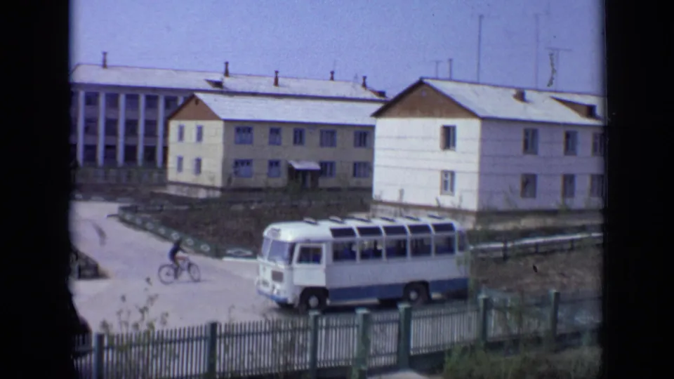 1971 Moscow street view with a white and blue bus, low residential buildings, fencing, and a cyclist passing nearby.
