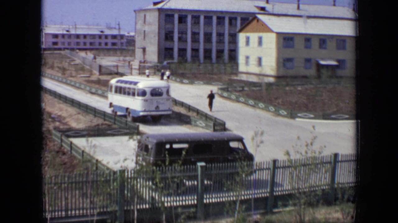 White and blue bus on a paved road beside pedestrians, fencing, and large buildings.
