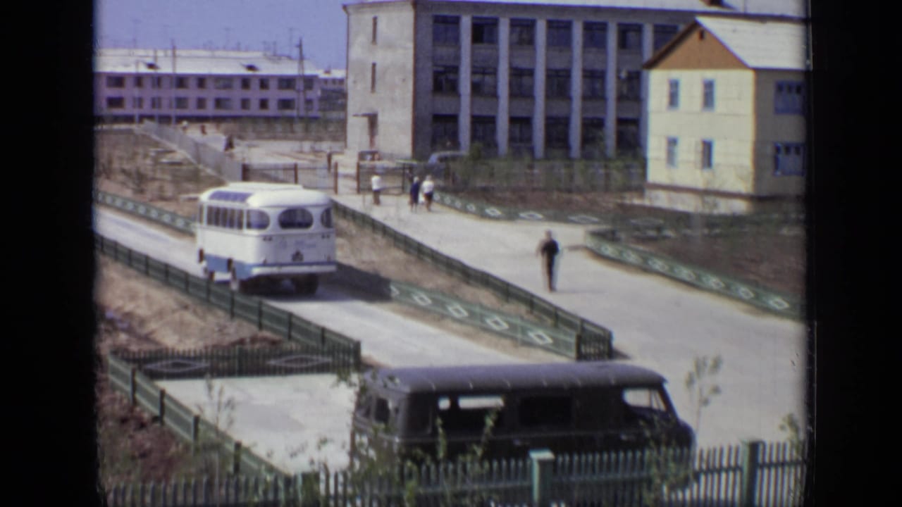 Bus on a quiet road near a fenced sidewalk, pedestrians, and block-like buildings.