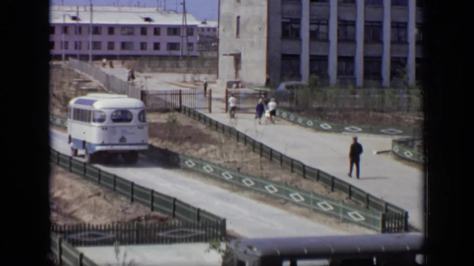 A Moscow road scene from 1971 with a bus, a dark vehicle in the foreground, pedestrians near a gate, and a lone man in dark clothing.