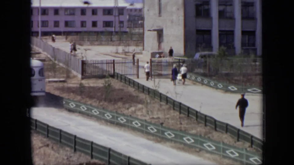 Late in the 1971 Moscow clip, pedestrians pass through or near a gate while the bus leaves the edge of the frame beside green fencing.