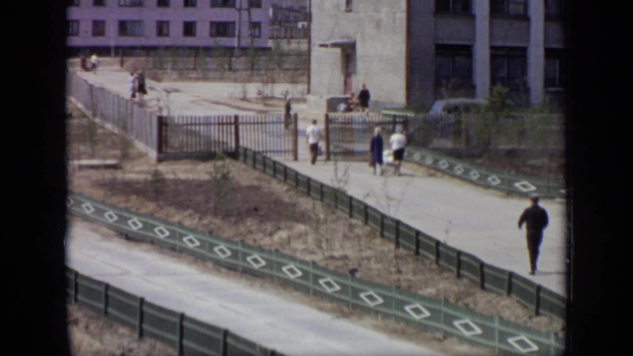 Pedestrians walking along a paved path beside green railings and apartment buildings.