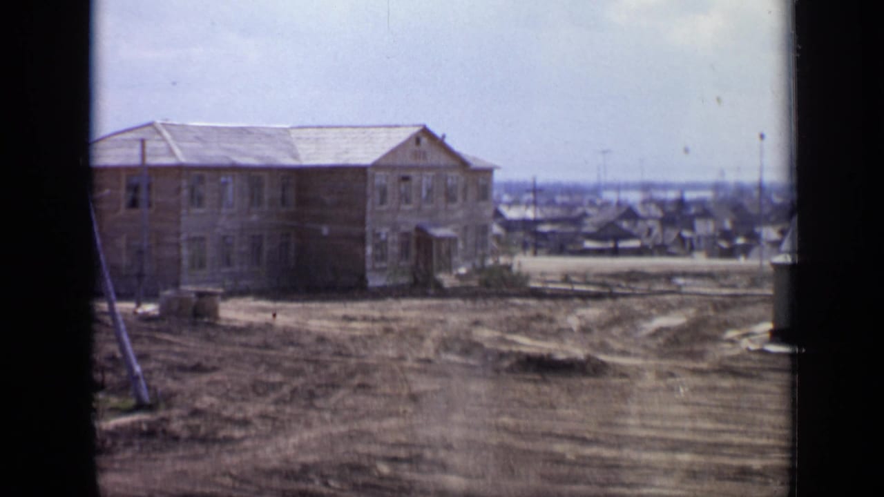 1971 Moscow outskirts view with a wooden building, uneven dirt roadway, low houses, and utility poles under a pale sky.