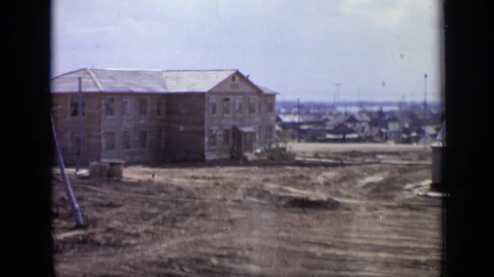 1971 Moscow outskirts view with a wooden building, uneven dirt roadway, low houses, and utility poles under a pale sky.
