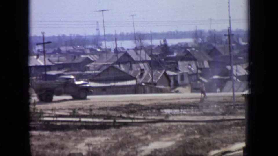 1971 Moscow roadside scene showing a dark pickup truck moving along a dusty road past low houses, utility poles, and a distant pedestrian.