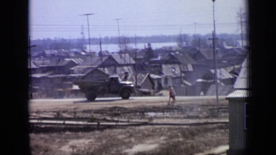 1971 Moscow home-movie frame with a pickup truck on a dirt road, a pedestrian nearby, low rooftops, and utility poles in the distance.