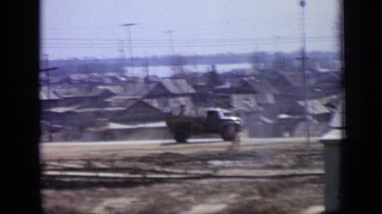 1971 Moscow residential roadside scene with a dark truck, low houses, a pedestrian and utility poles, seen through soft 8mm film.