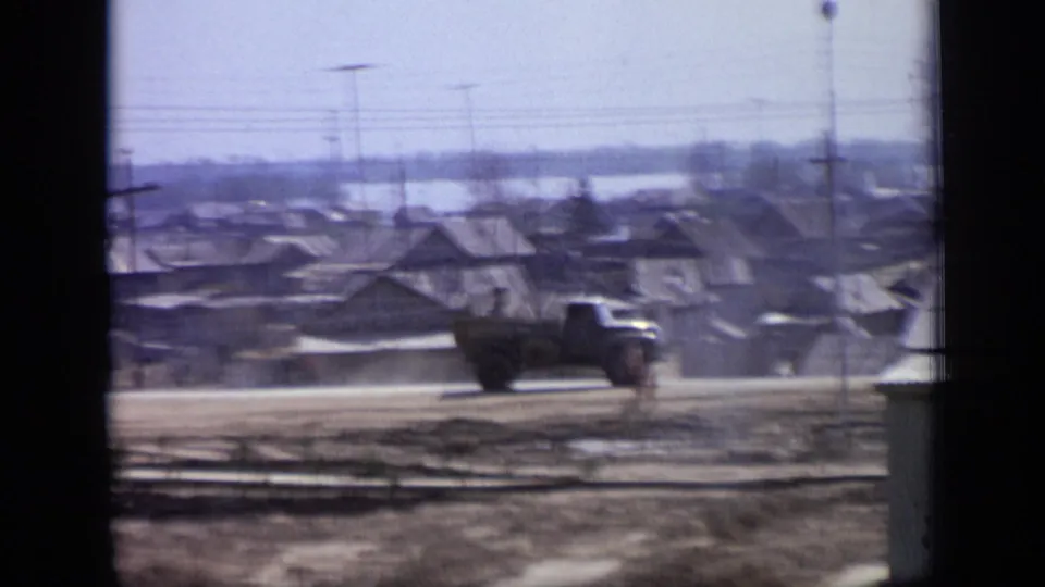 1971 Moscow residential roadside scene with a dark truck, low houses, a pedestrian and utility poles, seen through soft 8mm film.