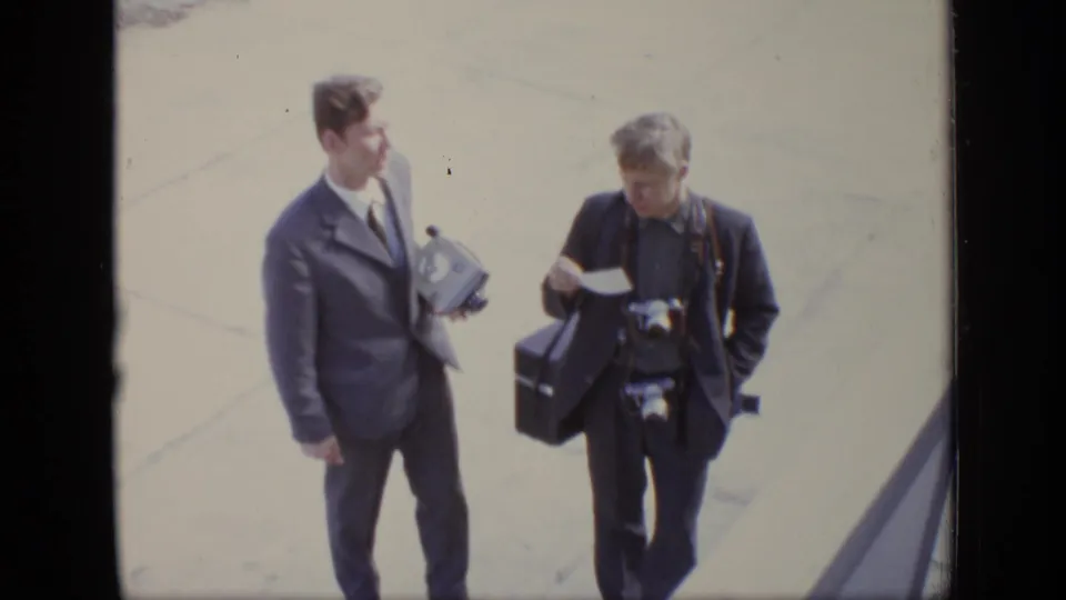 Two adult men walk across a paved area in Moscow, one with cameras around his neck and another holding a case and paper.