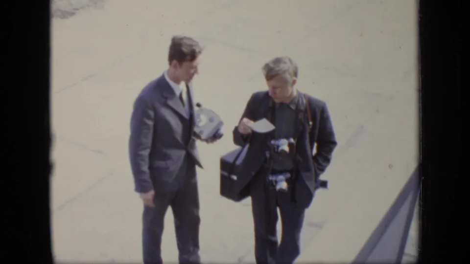 Two men in formal or dark clothing walk on pavement in the 1971 Moscow home-movie footage, carrying camera equipment and documents.