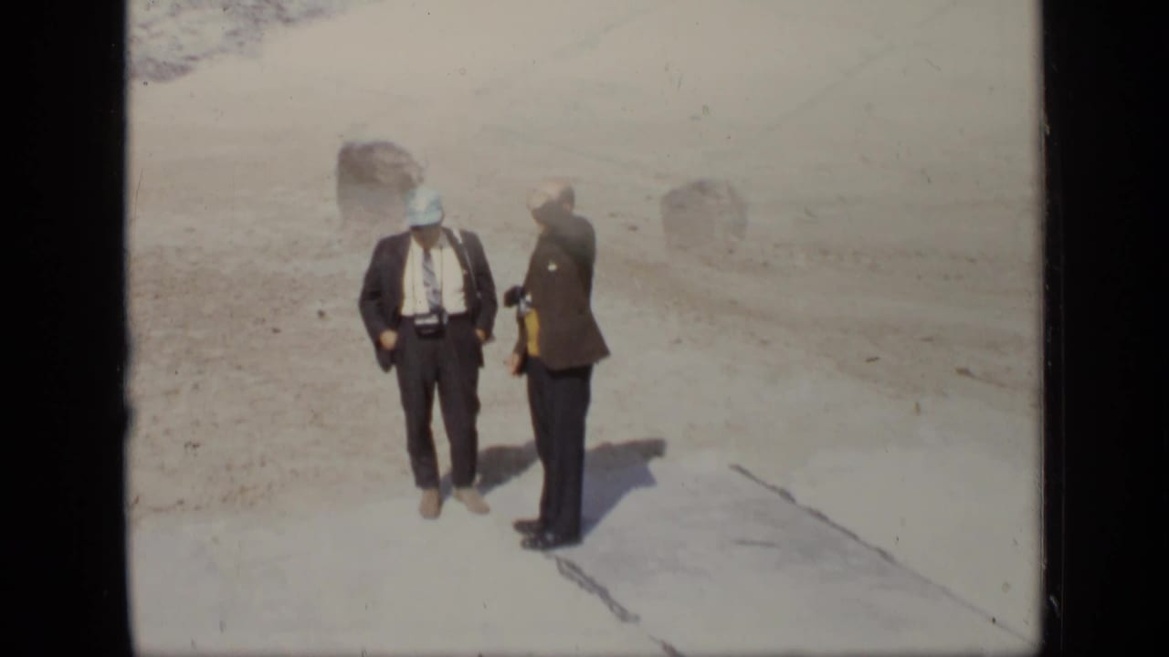 Two suited men stand on pale sandy ground at an outdoor site.