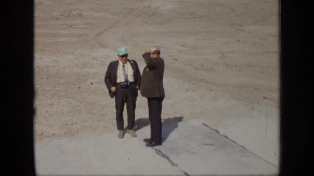 Two men stand together on a cleared outdoor site, one raising a hand to his face.