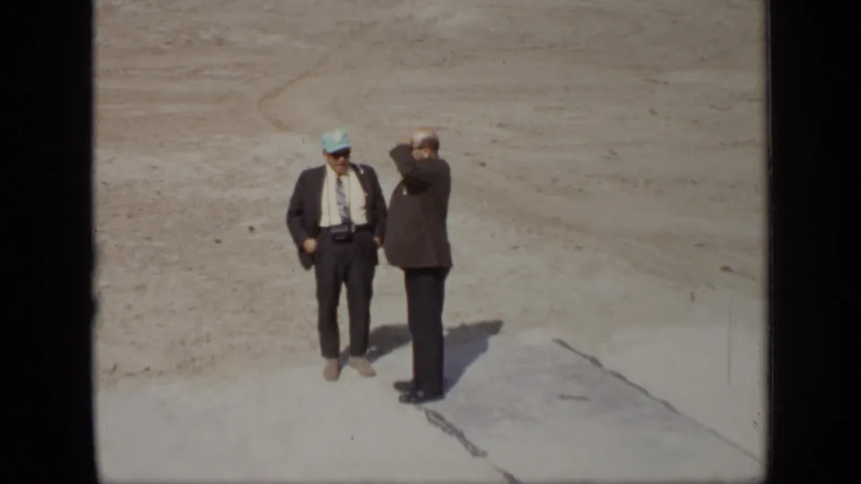 At the Moscow site, two suited men stand on open sandy ground while one raises a hand toward his face and the other faces the camera.
