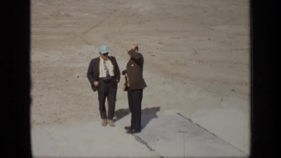 Two men in suits are seen from a slightly elevated 8mm viewpoint on the cleared Moscow site, with equipment visible at the waist of one man.