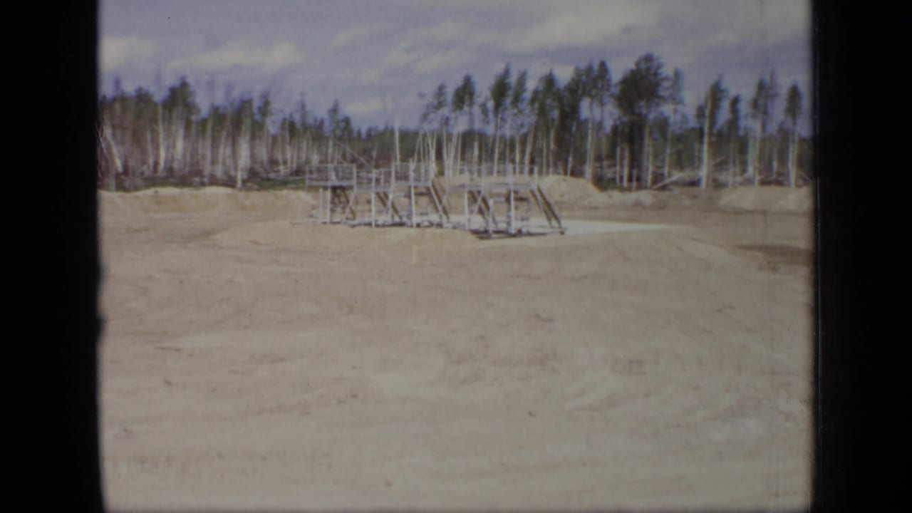 Cleared sandy ground with a small metal platform structure and birch trees behind it.