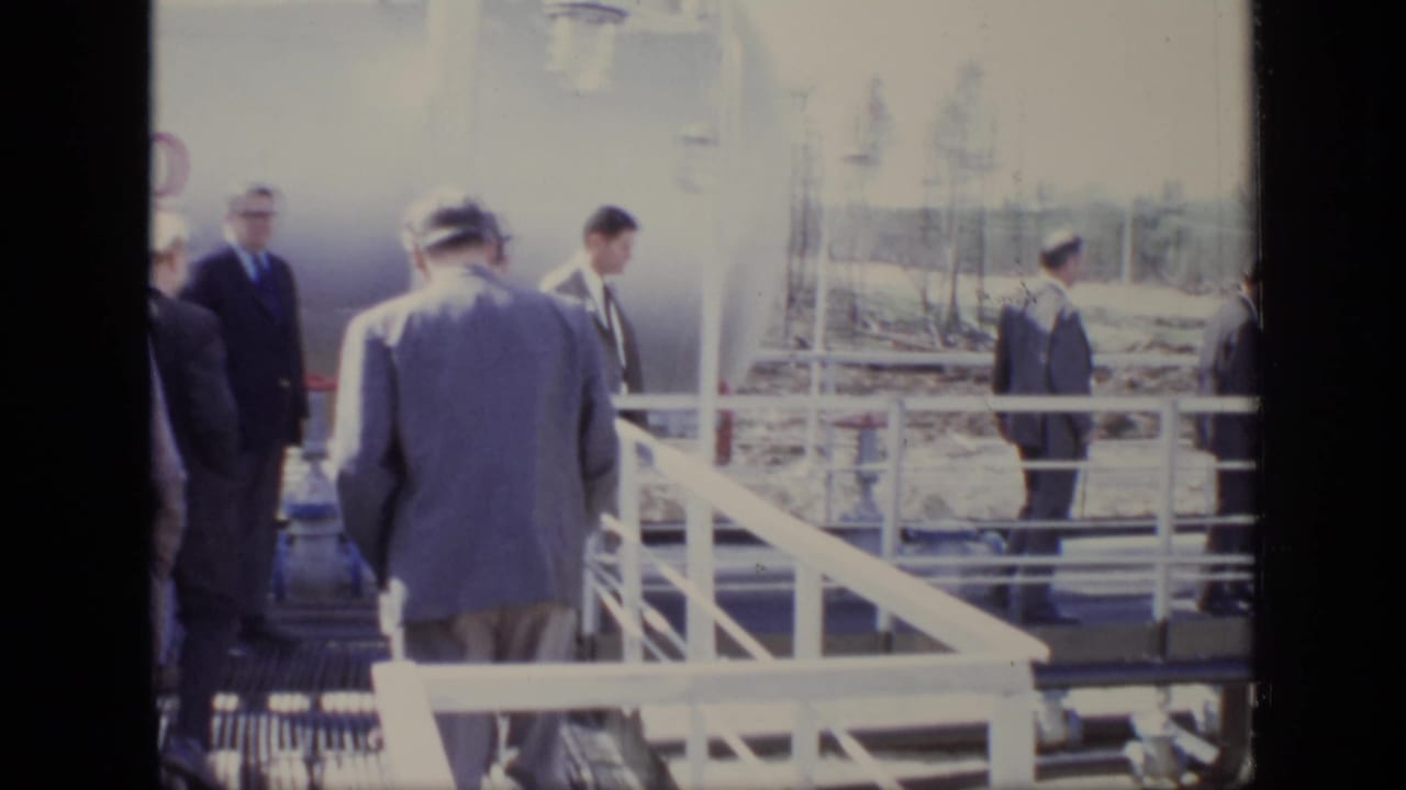 In 1971 Moscow footage, several suited men move along a raised platform beside large tanks, white railings, and pipework.