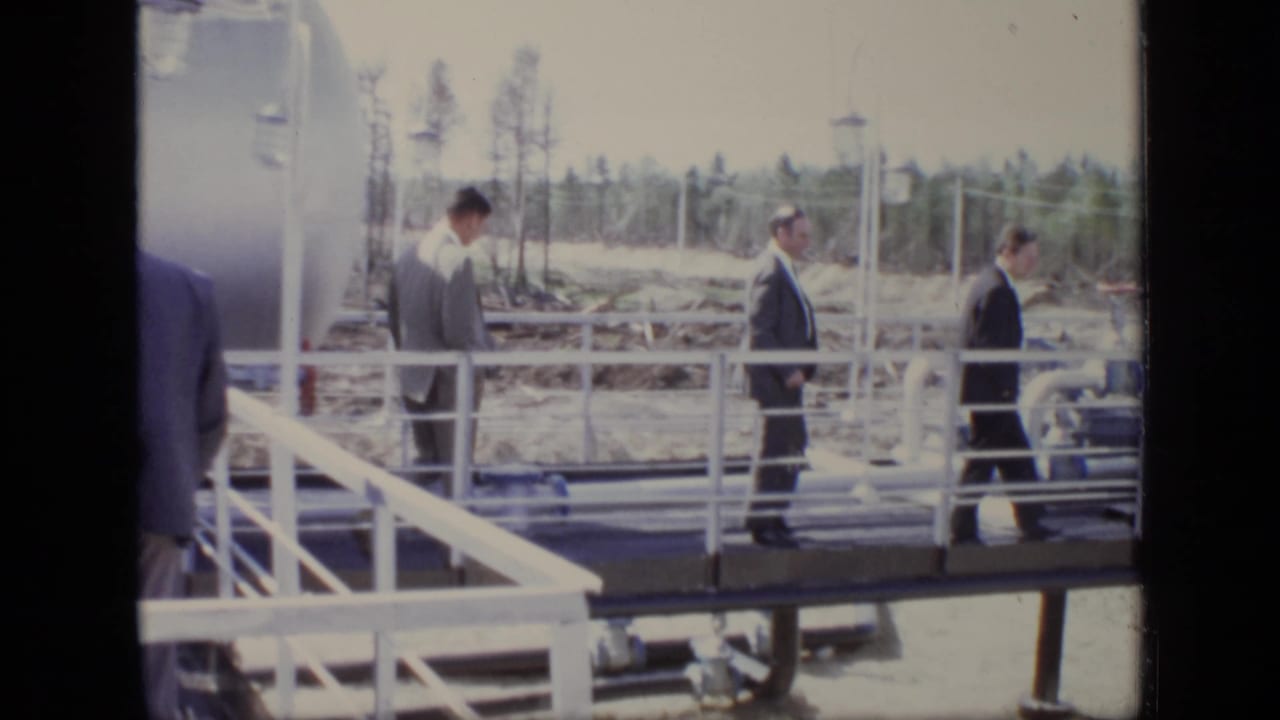 Suited men continue across a white-railed platform at a Moscow industrial site, with tanks, pipes, and cleared ground visible.