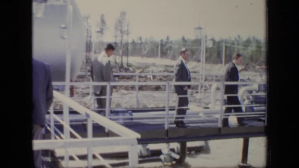Suited men continue across a white-railed platform at a Moscow industrial site, with tanks, pipes, and cleared ground visible.