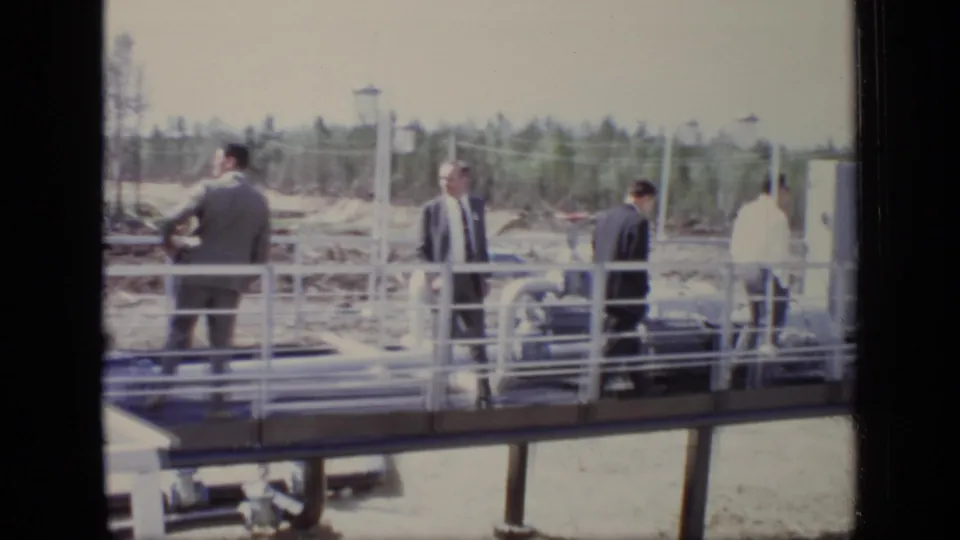 The 1971 Moscow scene shows men in suits on a raised platform near exposed pipes, railings, cleared earth, and distant trees.