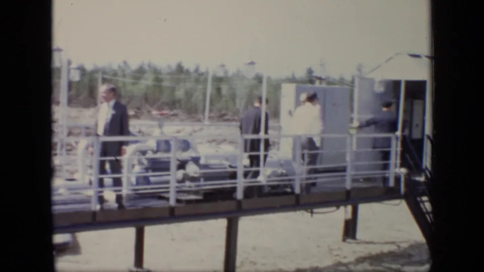 At the Moscow industrial site, suited men gather near a small white shed, white railings, pipes, and a raised platform.