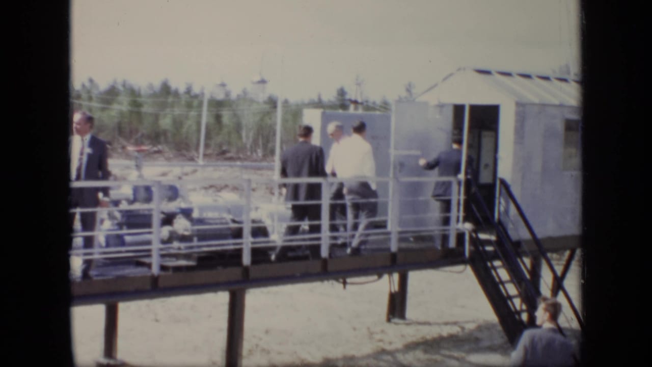 Men in suits stand on a raised wooden walkway beside a small structure and railings.