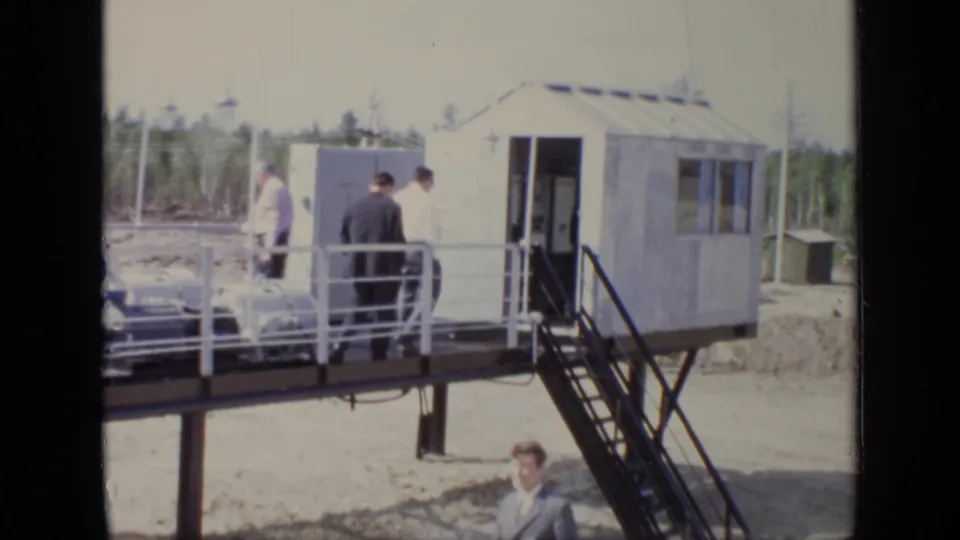 Suited men move along a raised platform at a Moscow industrial site in 1971, with stairs, railings, and a small structure visible.