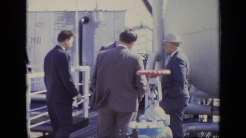 At the industrial site, men in suits and a hard hat examine equipment beside a white tank, pipes, railings, and a red valve.